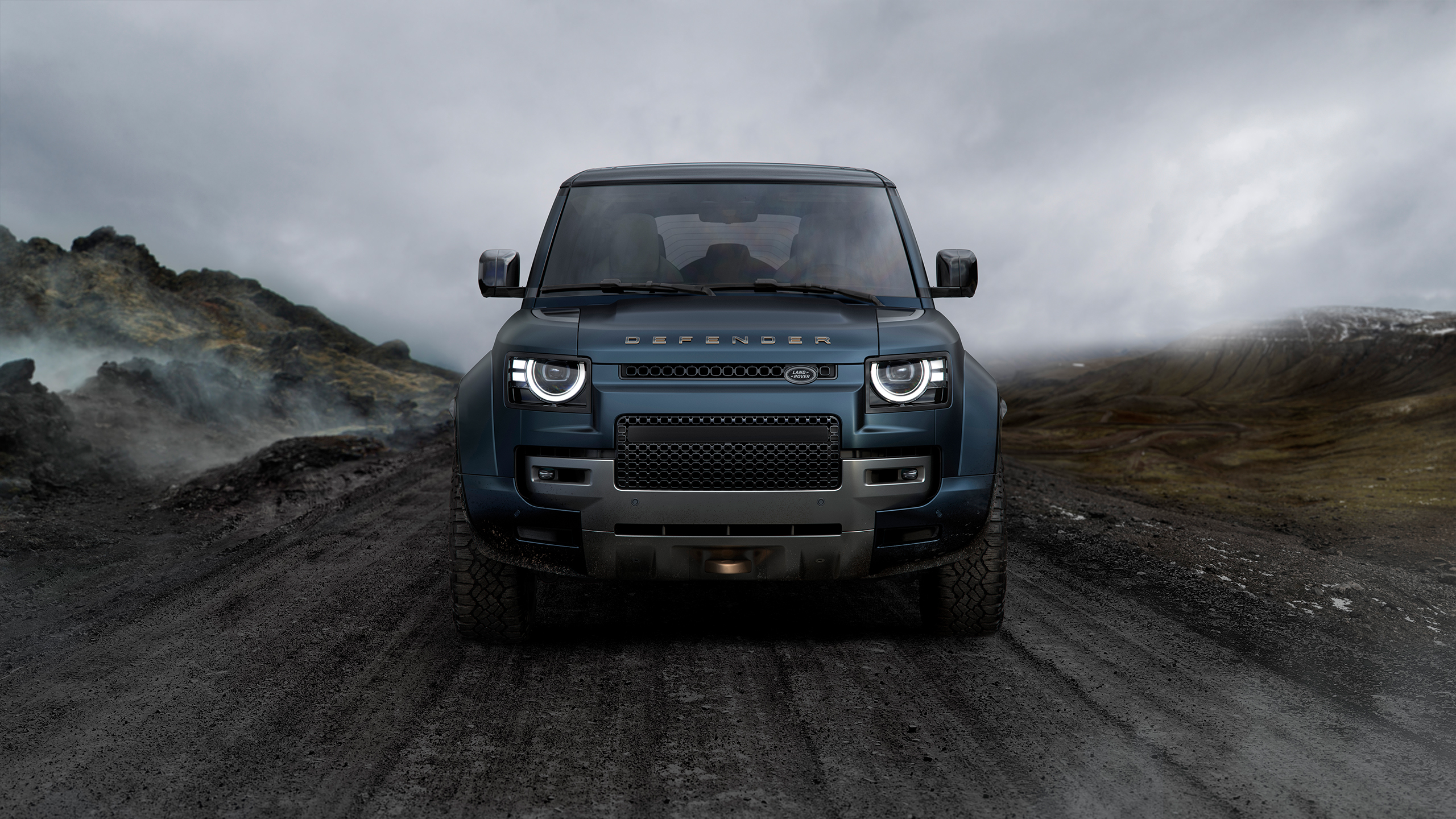 Front view of a blue Defender on a rugged mountain road under cloudy skies.
